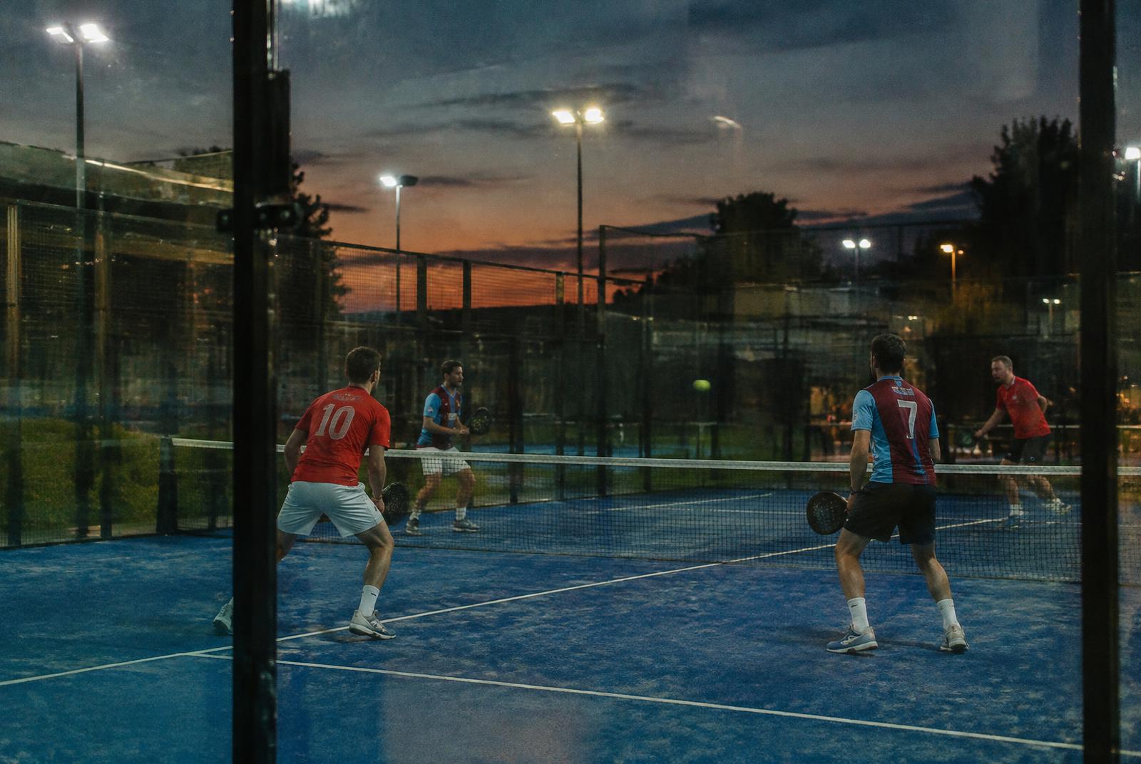 Rival football supporters in club shirts playing doubles padel under floodlights